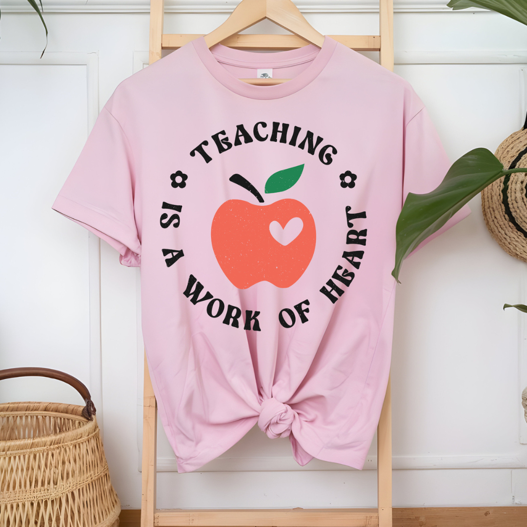 Light pink graphic teacher tee featuring an apple with a heart and the phrase "Teaching Is a Work of Heart."