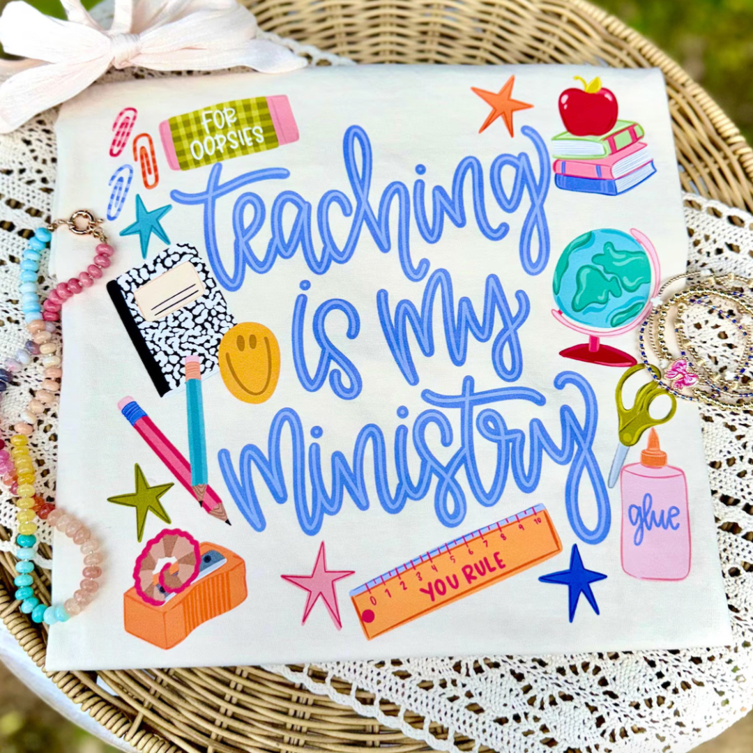 White teacher graphic tee featuring colorful classroom supplies surrounding the phrase "Teaching Is My Ministry."