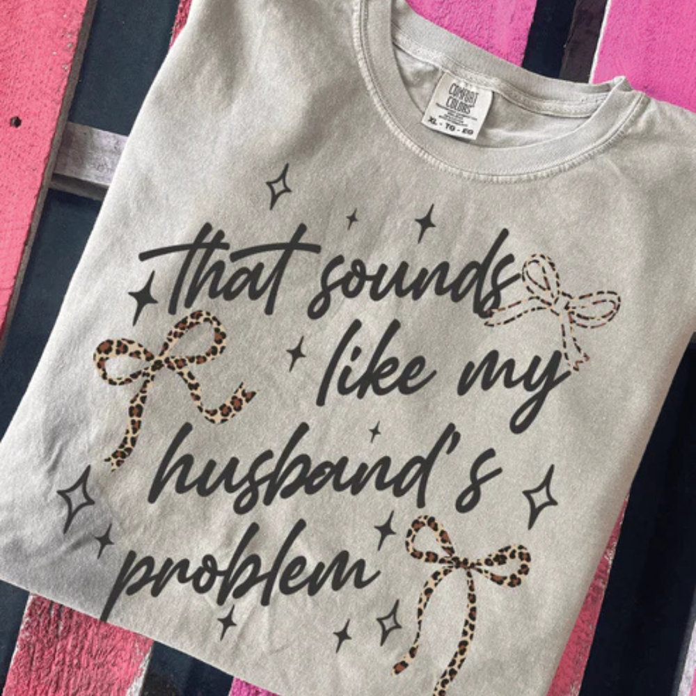 Cream graphic t-shirt featuring the phrase That Sounds Like My Husband's Problem with leopard print bows and whimsical sparkle details.