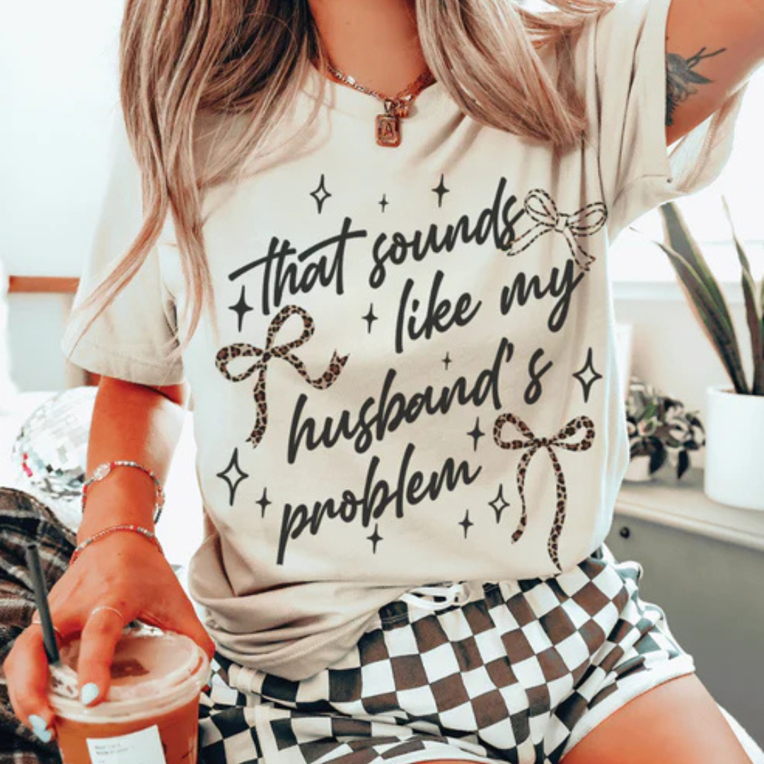 Cream graphic t-shirt featuring the phrase That Sounds Like My Husband's Problem with leopard print bows and whimsical sparkle details.
