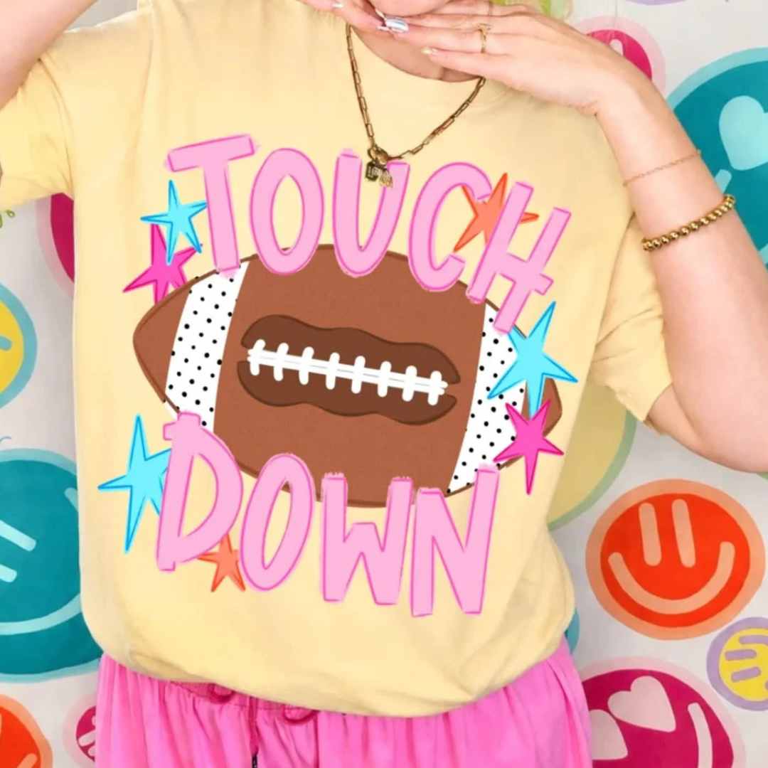 Butter yellow graphic tee featuring oversized pink "Touch Down" lettering over a football with colorful star accents.