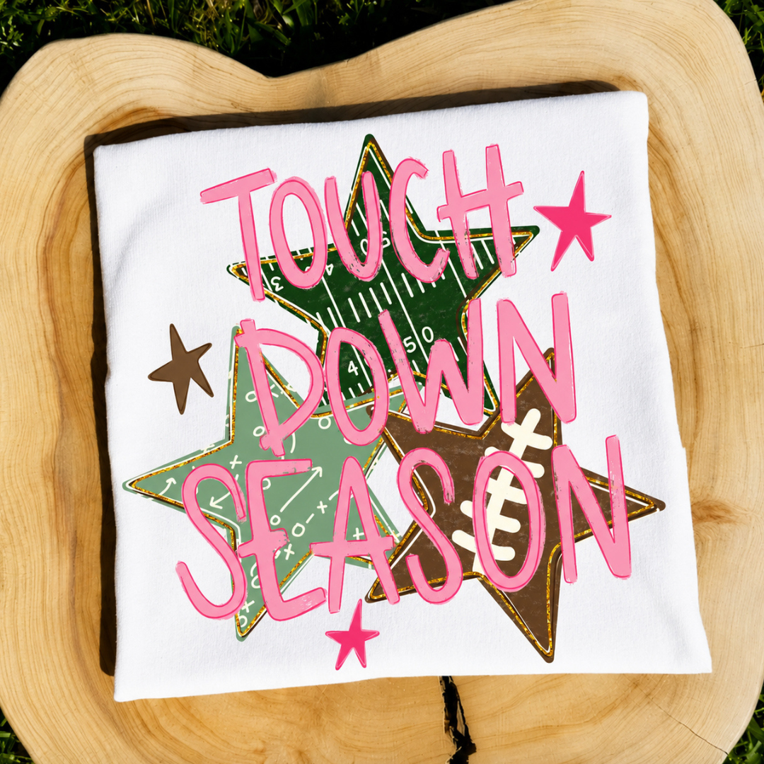 White graphic tee featuring bright pink "Touch Down Season" lettering over football playbook-inspired star graphics.