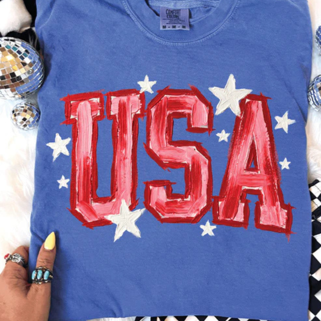 Flo blue graphic tee featuring oversized red USA lettering with white star accents in a patriotic design.