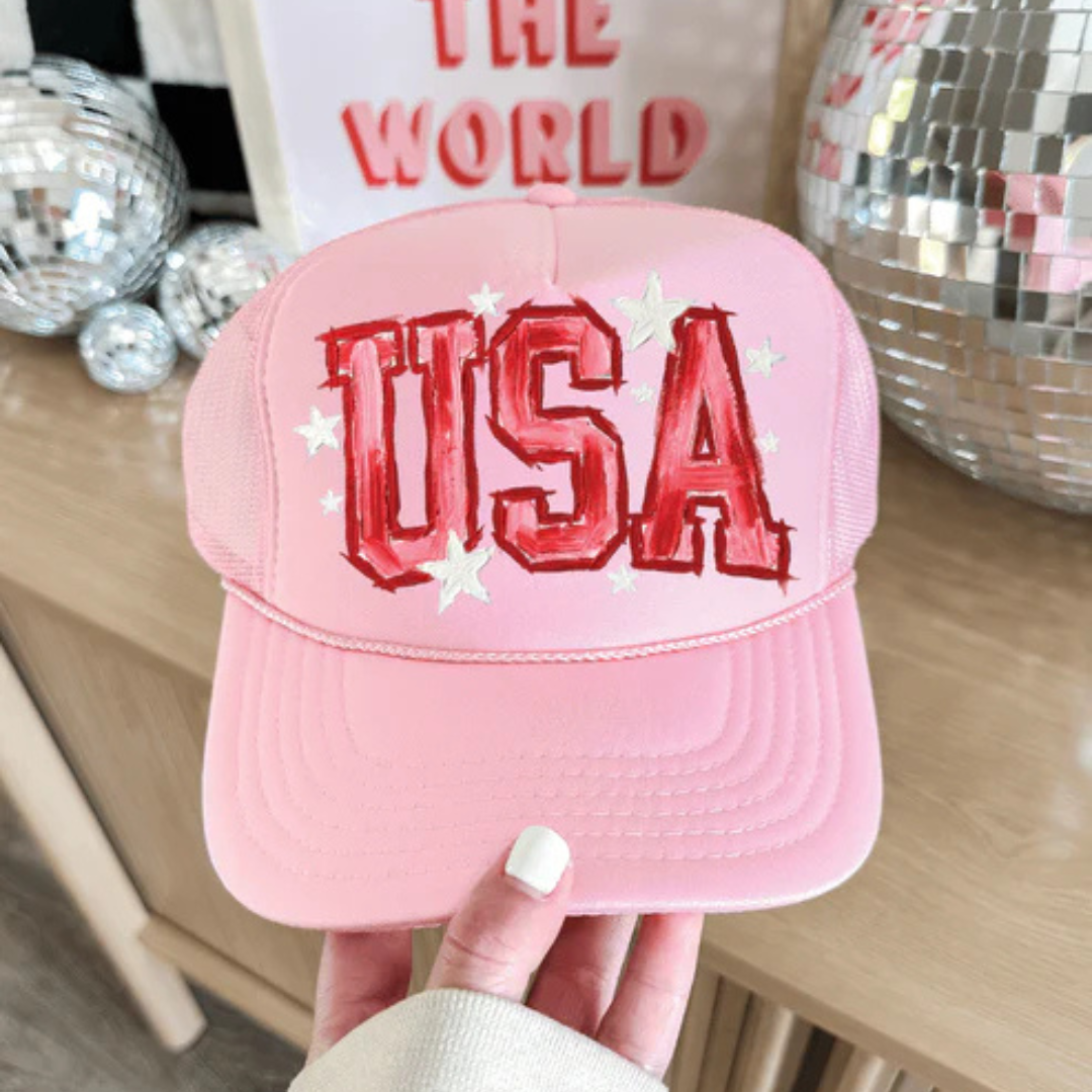 Baby pink foam trucker hat featuring oversized USA lettering with white star accents in a patriotic design.