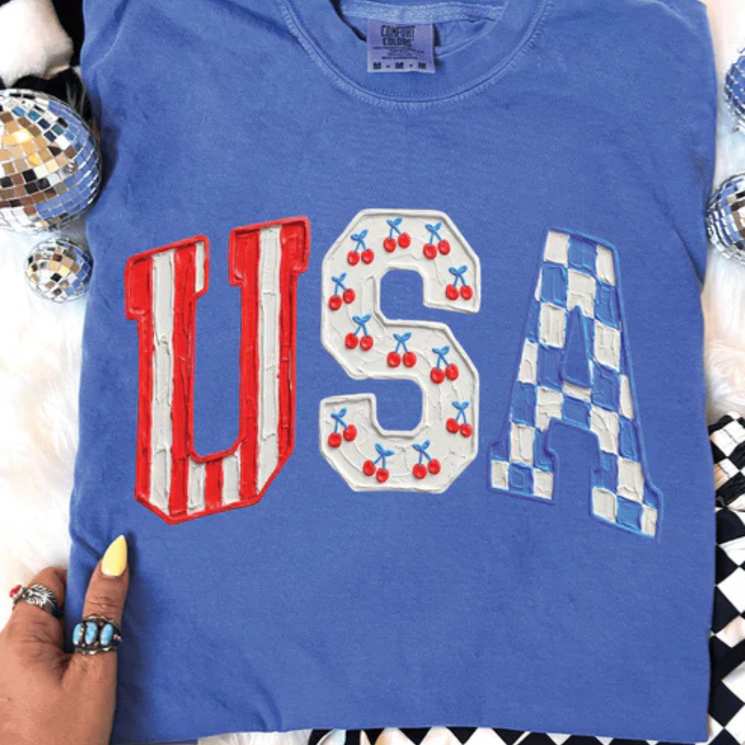 Flo blue graphic tee featuring oversized USA lettering with cherry and blue checkered patriotic accents.
