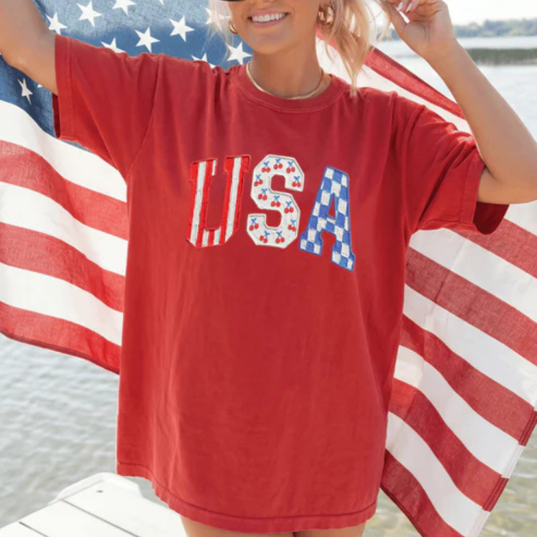 Red graphic tee featuring oversized USA lettering with cherry and blue checkered patriotic accents.
