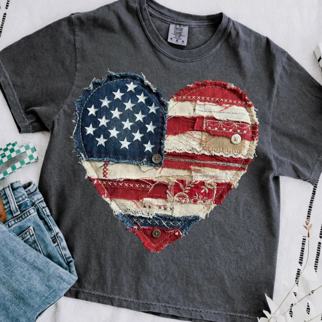 Washed charcoal gray graphic tee featuring a vintage patchwork American flag heart with distressed denim, lace, button, and stitched details.
