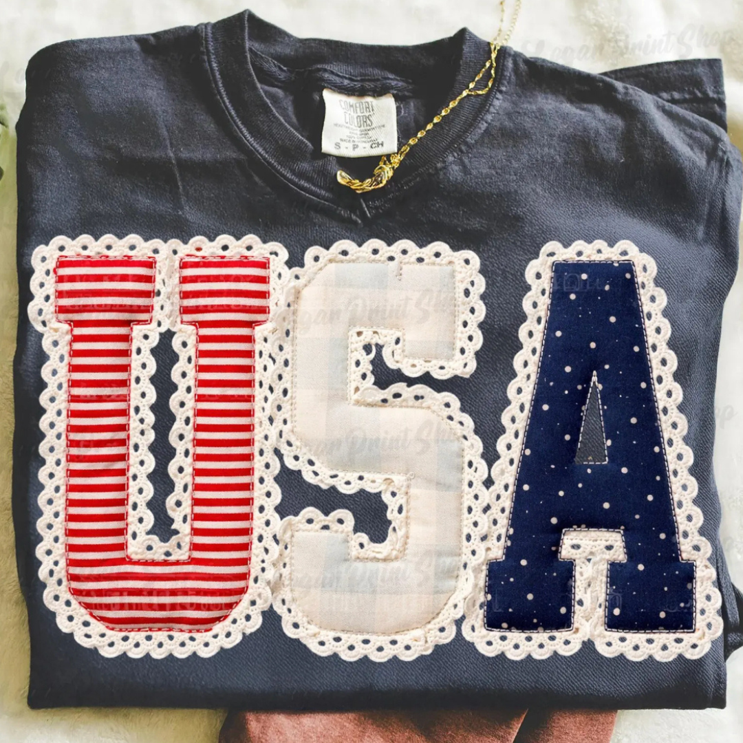 Navy graphic tee featuring oversized USA lettering with red and white stripes, neutral plaid, navy polka dots, and eyelet lace trim details.