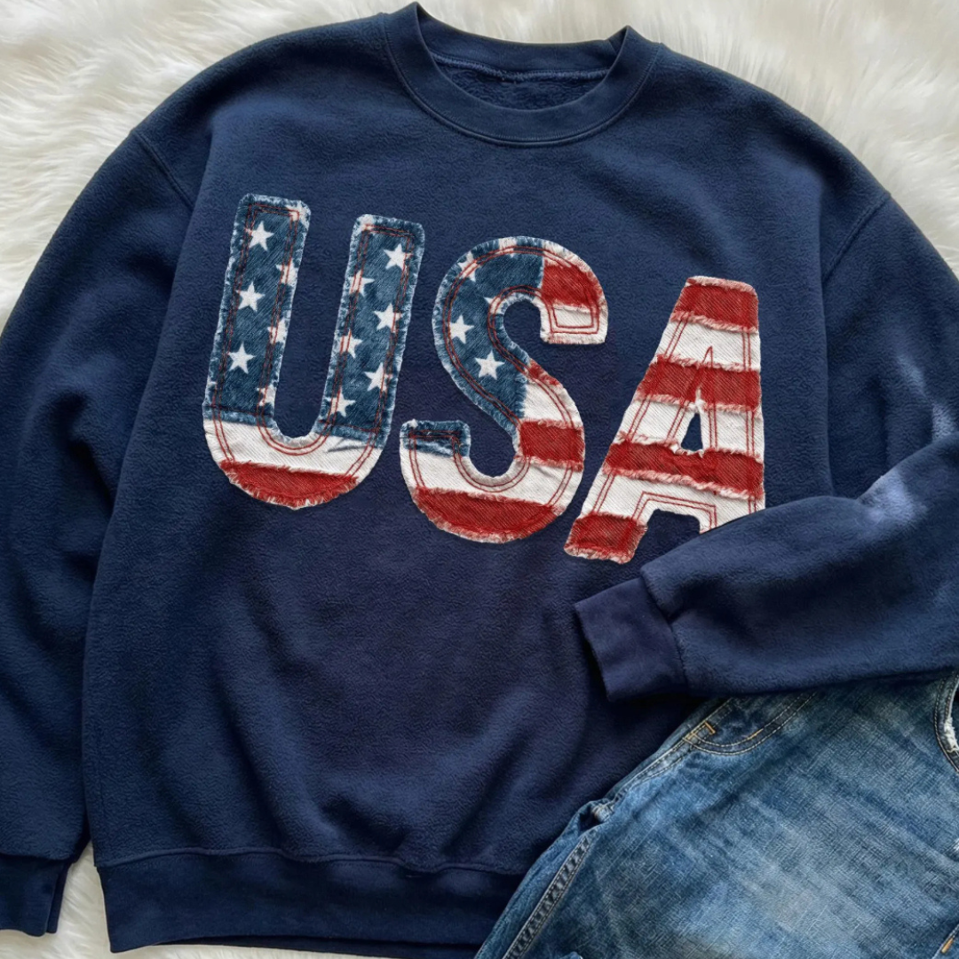 Navy patriotic sweatshirt featuring oversized USA lettering with distressed American flag patchwork artwork and vintage Americana styling.