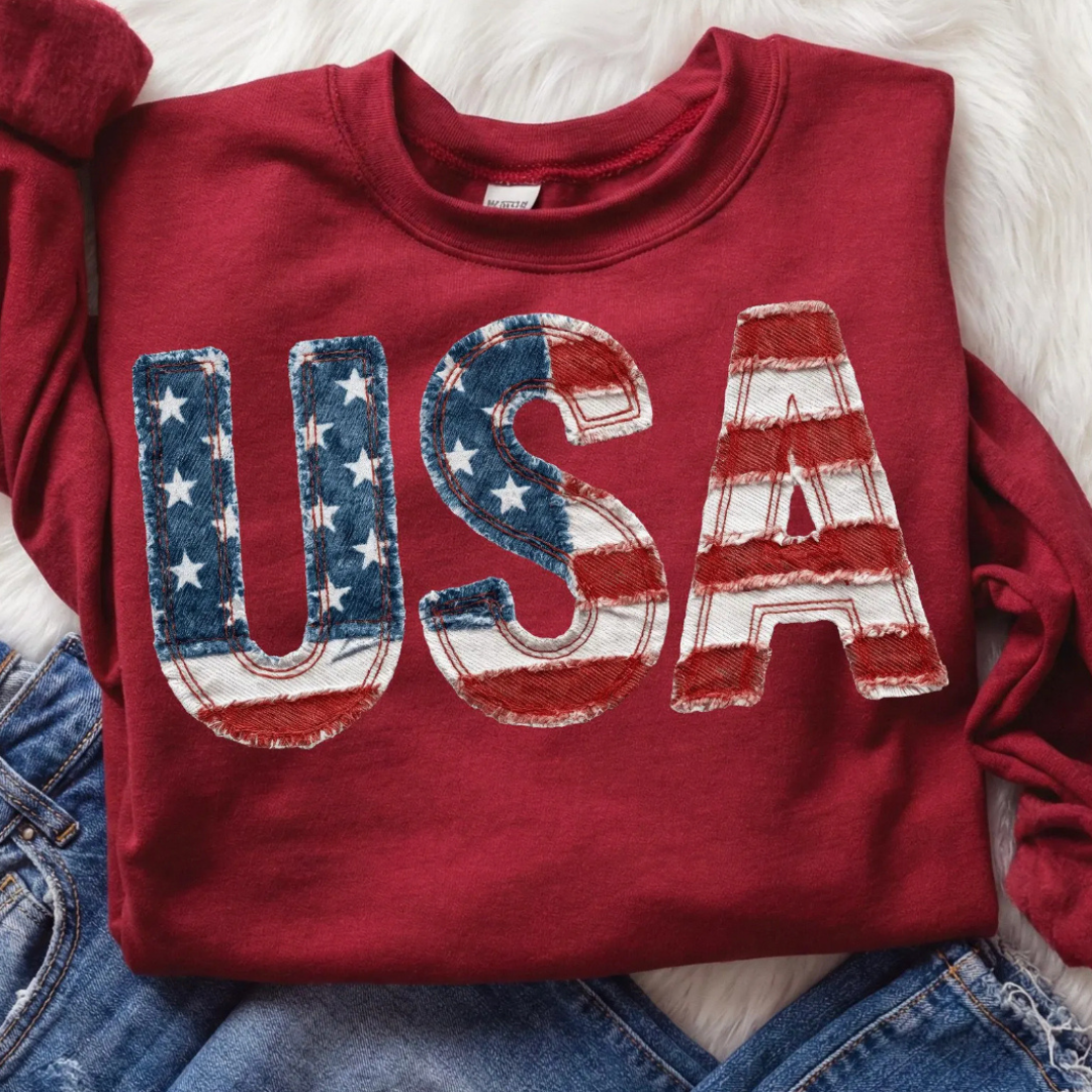 Red patriotic sweatshirt featuring oversized USA lettering with distressed American flag patchwork artwork and vintage Americana styling.