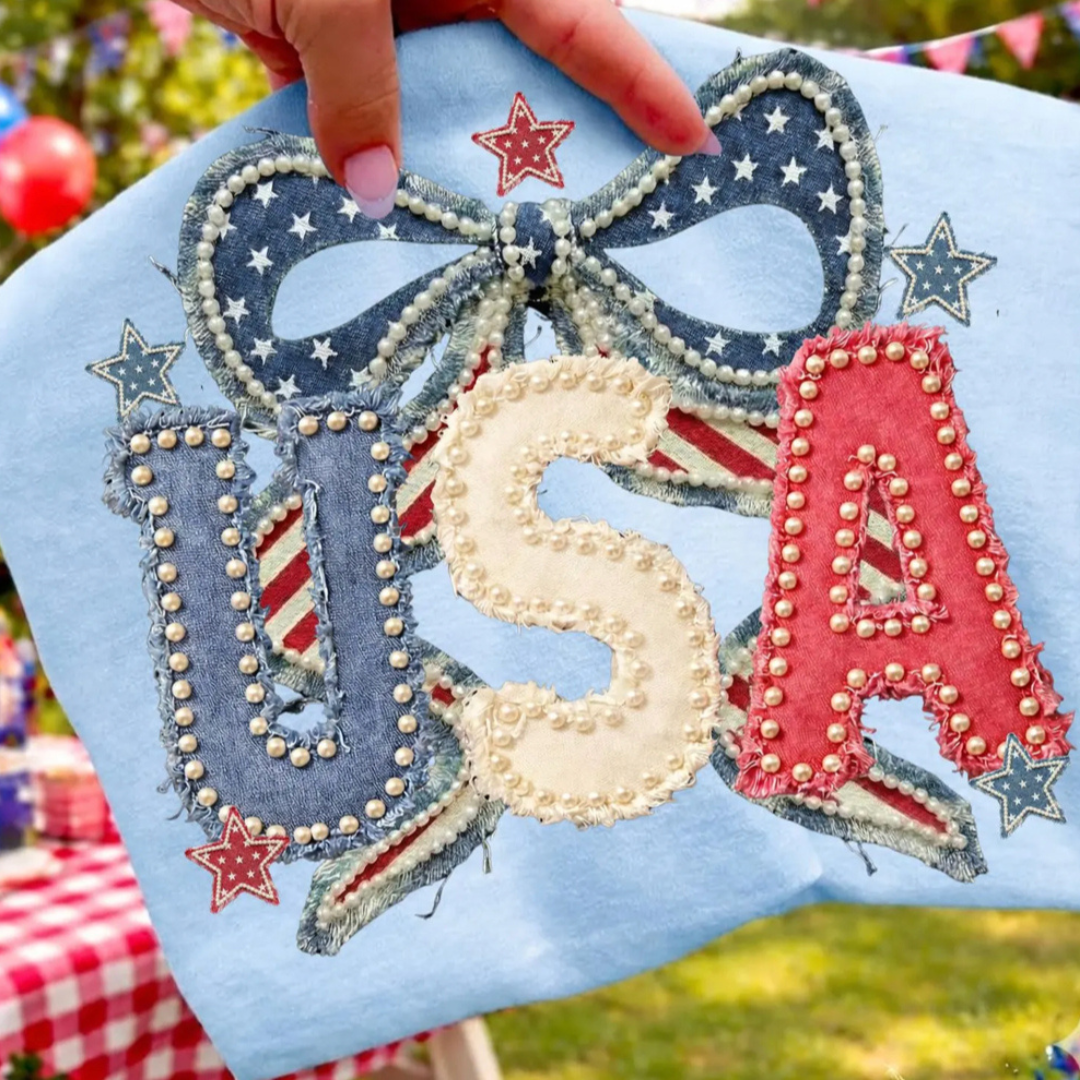 Light blue graphic tee featuring pearl-trimmed USA lettering with a patriotic denim bow, stars, and red, white, and blue Americana details.