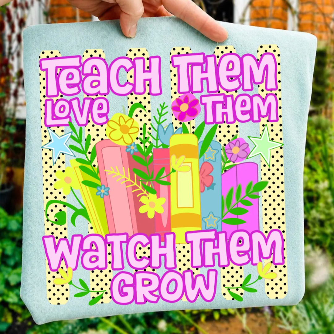 Light blue teacher graphic tee featuring colorful books, flowers, and the phrase "Teach Them, Love Them, Watch Them Grow" over a playful polka dot background.