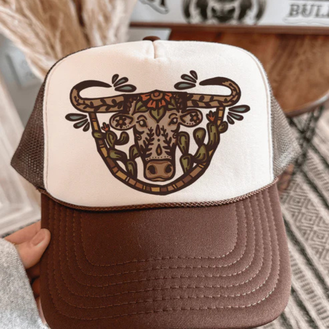 Brown and cream foam trucker hat featuring an artistic western cattle design with floral and cactus details and adjustable snapback closure.