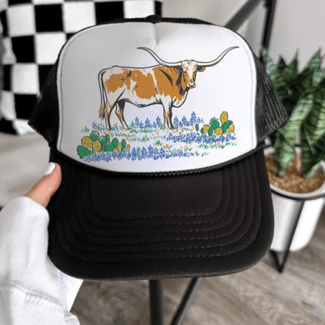 Trucker hat with a cow and cactus design held by a hand in a casual setting.
