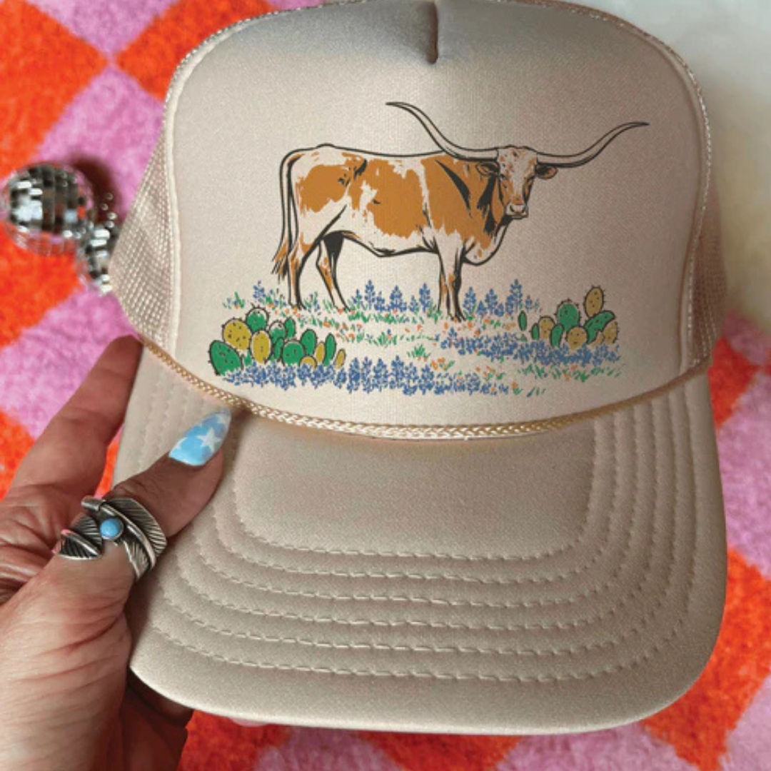 Beige cap with a cow and floral design held by a hand on a checkered background