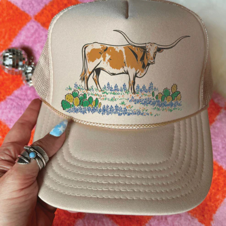 Beige cap with a cow and floral design held by a hand on a checkered background