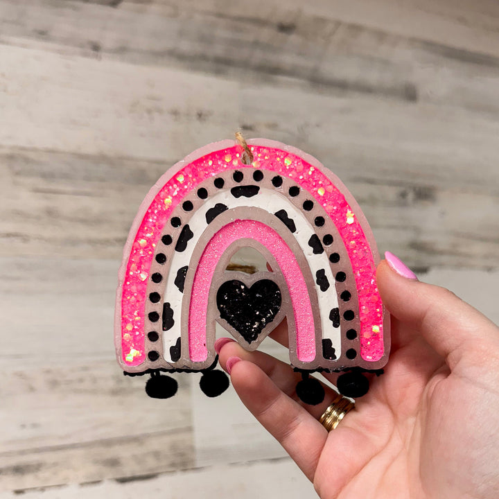 A wooden background with a rainbow wax car freshie in black and pink. A heart in the center with black fringe at the end of the rainbow.