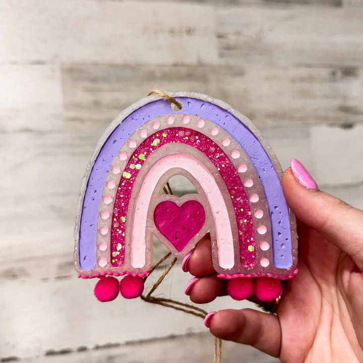 A wooden rustic background with pink and purple rainbow freshie with pom poms.