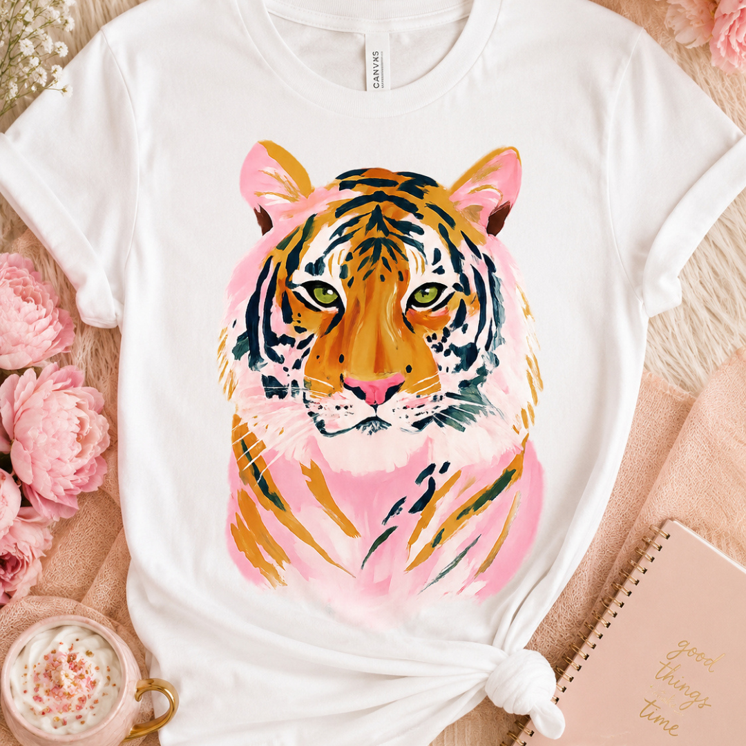 White graphic tee featuring a watercolor-style tiger design with pink accents, orange details, and green eyes.