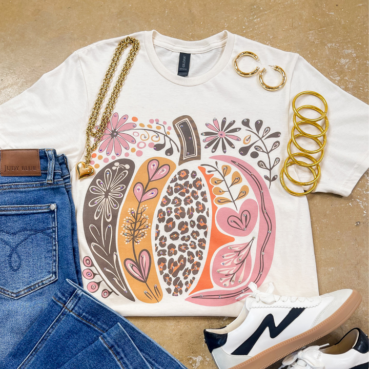 White t-shirt with floral and leopard print design, blue jeans, white sneakers, and gold jewelry on a beige surface.