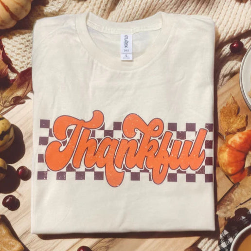 Folded white t-shirt with 'Thankful' design on a wooden surface with autumn decorations.