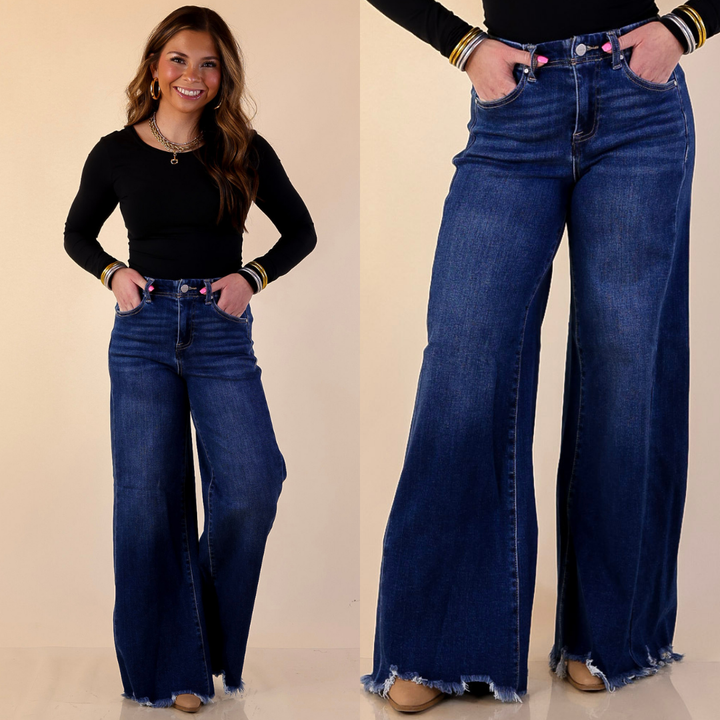 Model is wearing Risen Wide Leg Jeans with Raw Hem in dark wash with a black top against a beige background. 