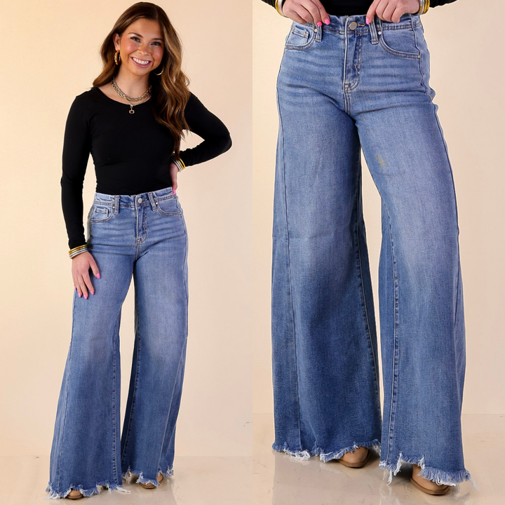 Model is wearing Risen WIde Leg Jeans with Raw Hem in Medium Wash with a black top against a beige background. 