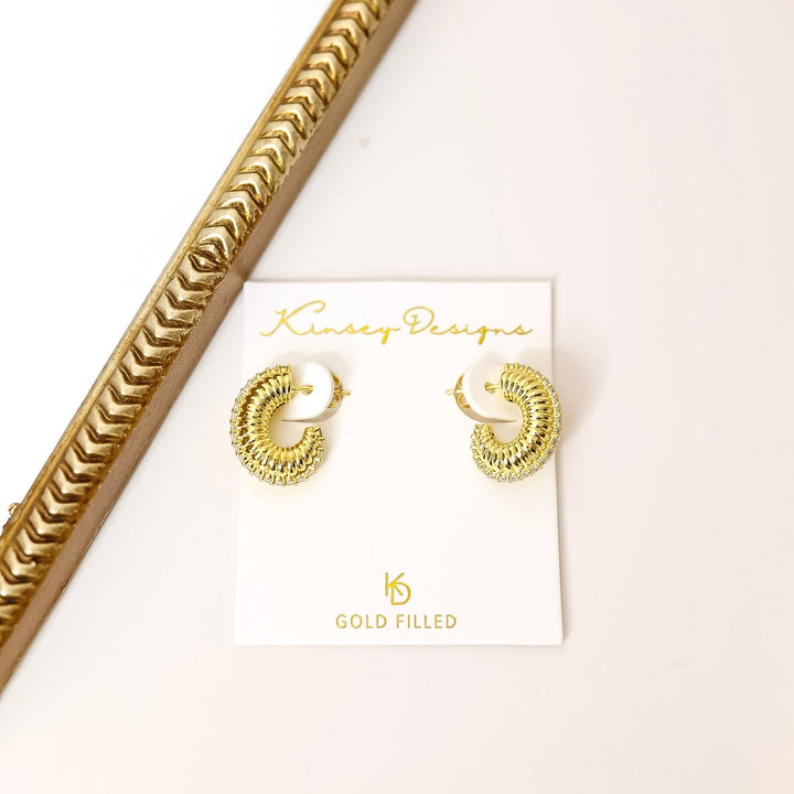 Gold earrings on a Kinsey Designs branded card with a gold frame in the background.