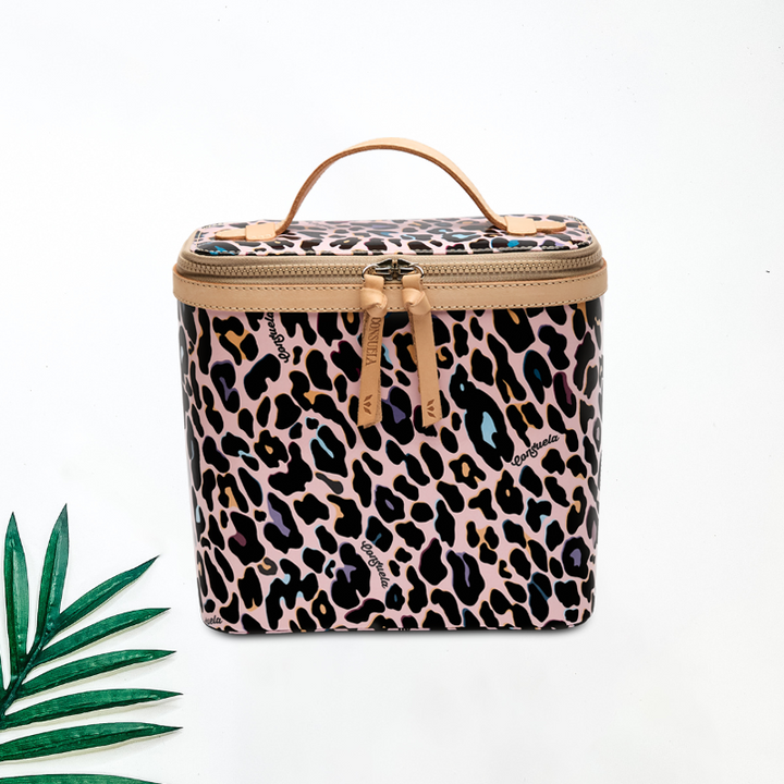A solid white background with a palm tree leaf. Consuela's slim train case in the print Ruth, a colorful jag print and genuine leather zipper plus handle combo on the exterior. The interior features a Vinyl material for easy clean and mesh pocket with two elastic makeup brush organizers