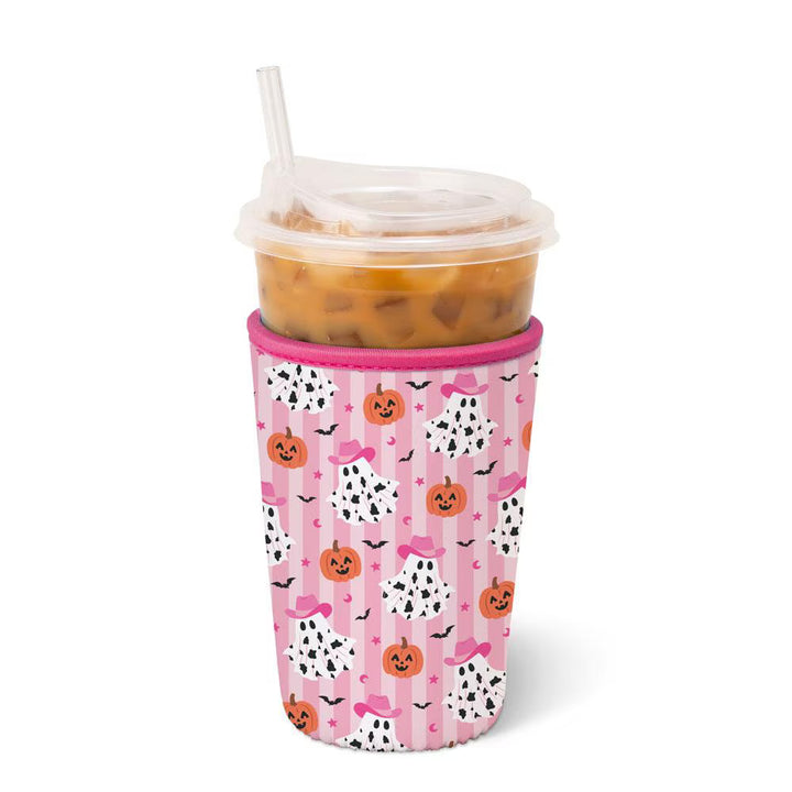 A pink insulated travel tumbler sleeve with a Halloween pattern of pumpkins, ghosts, bats, and hearts, shown on a clear plastic cup with a straw and iced drink against a white background.