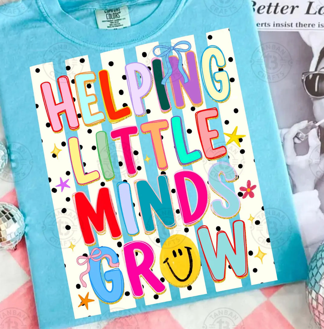 Turquoise blue graphic tee featuring the phrase "Helping Little Minds Grow" in colorful lettering with bows, smiley face, stars, and a black and white polka dot striped background.