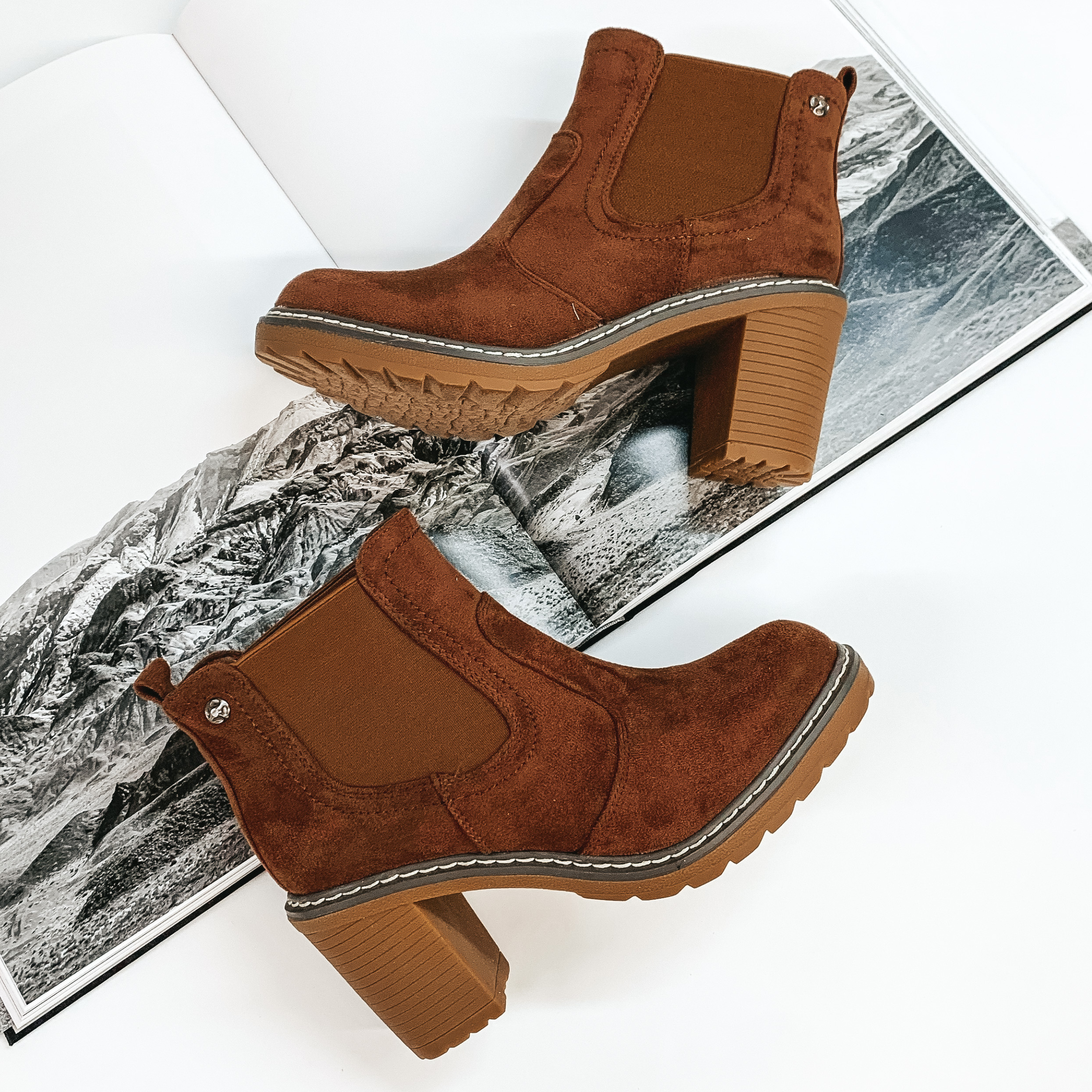 Brown suede booties with a tan rubber sole and chunky heel. These booties are pictured laying on an open book on a white background. 
