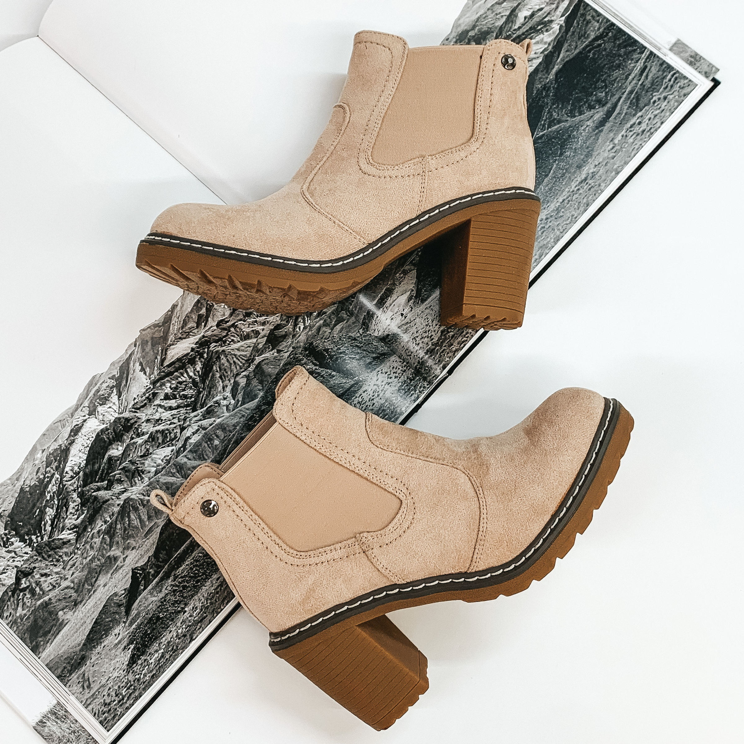 Tan suede booties with a tan rubber sole and chunky heel. These booties are pictured laying on an open book on a white background. 
