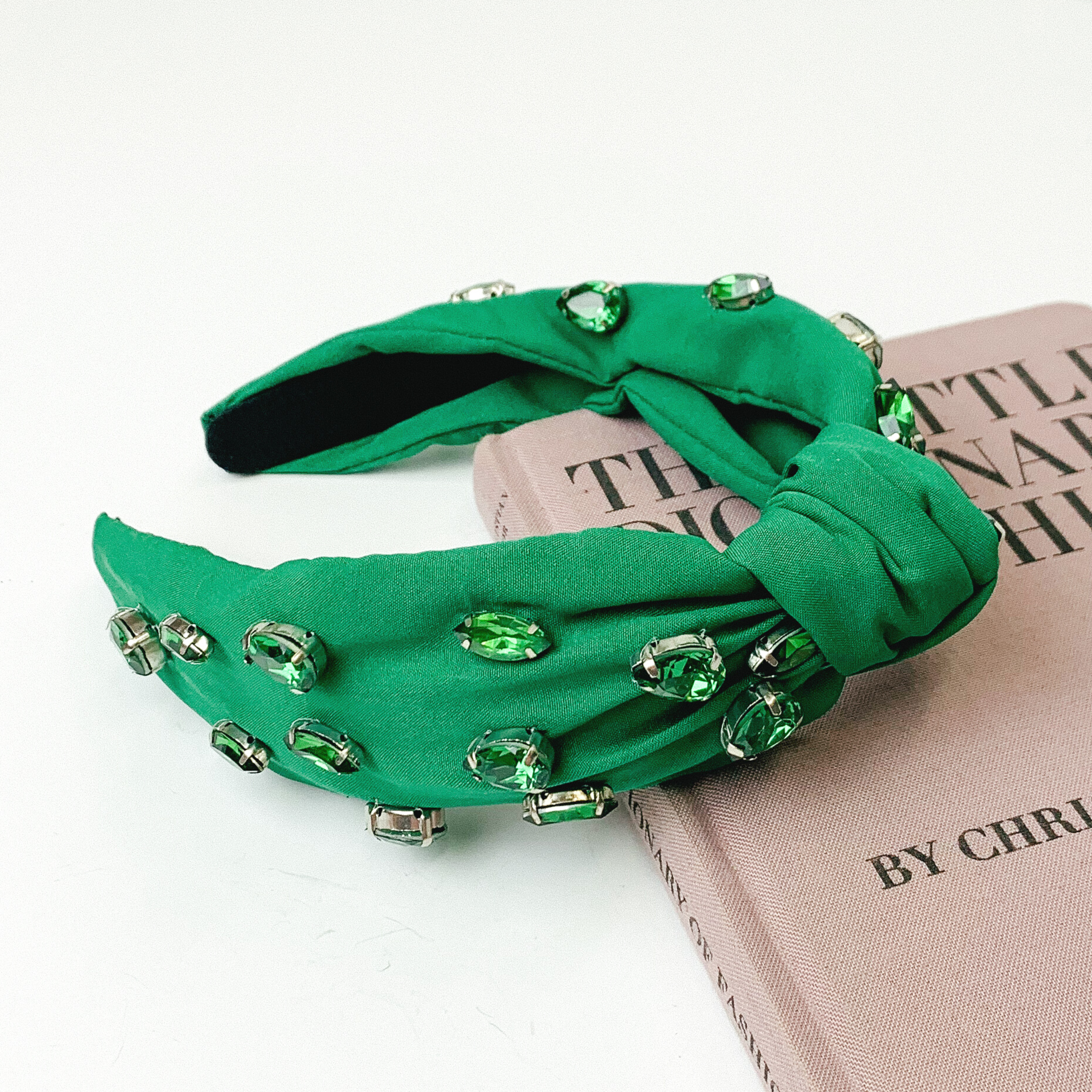 Green colored knot headband with different shaped green crystals. This headband is pictured partially laying on a mauve book on a white background.