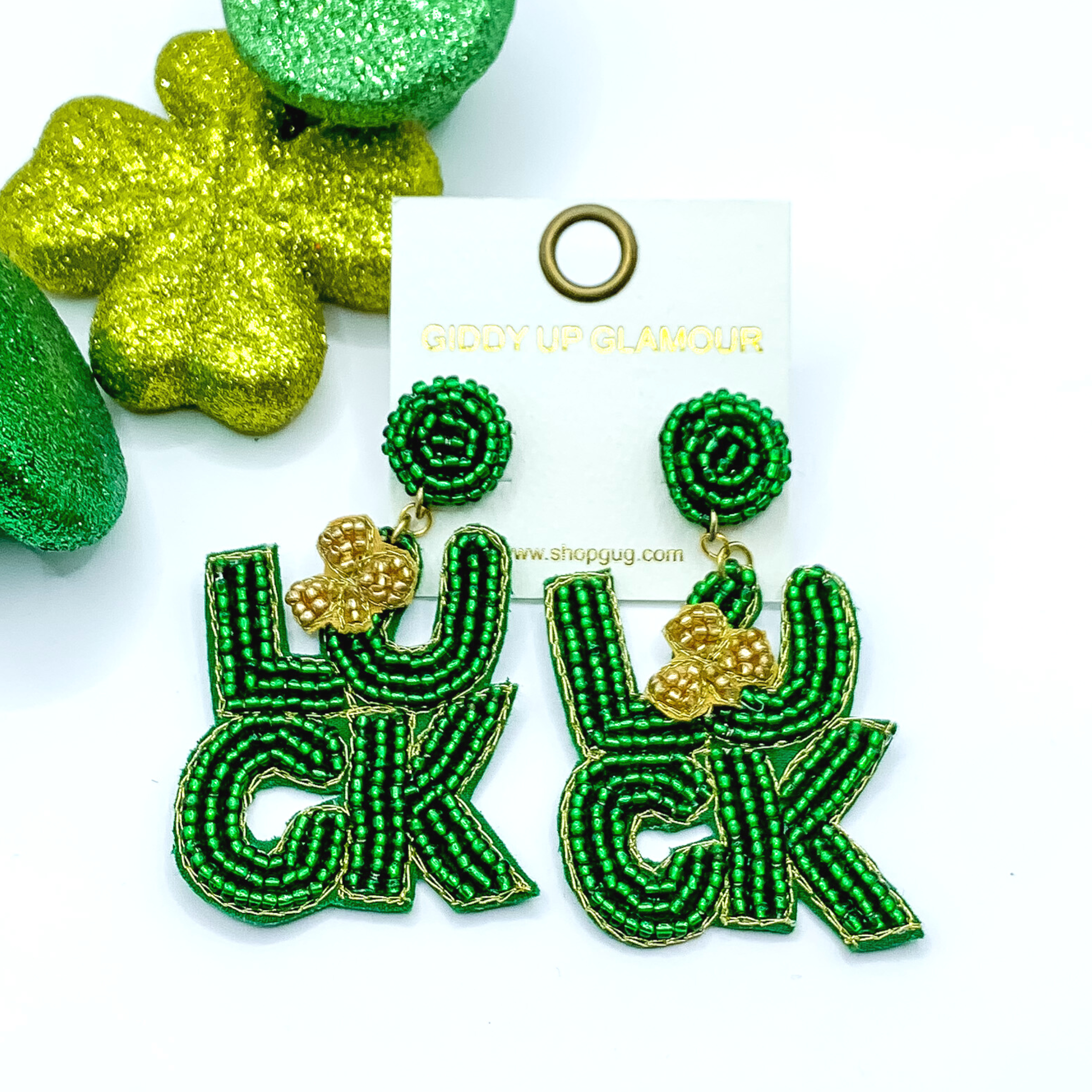 Green beaded circle post back earrings with a beaded pendant. The pendant is the word "LUCK" spelled out with green beads and includes a gold clover on the "U." These earrings are pictured on a white background with green decor in the top left corner. 