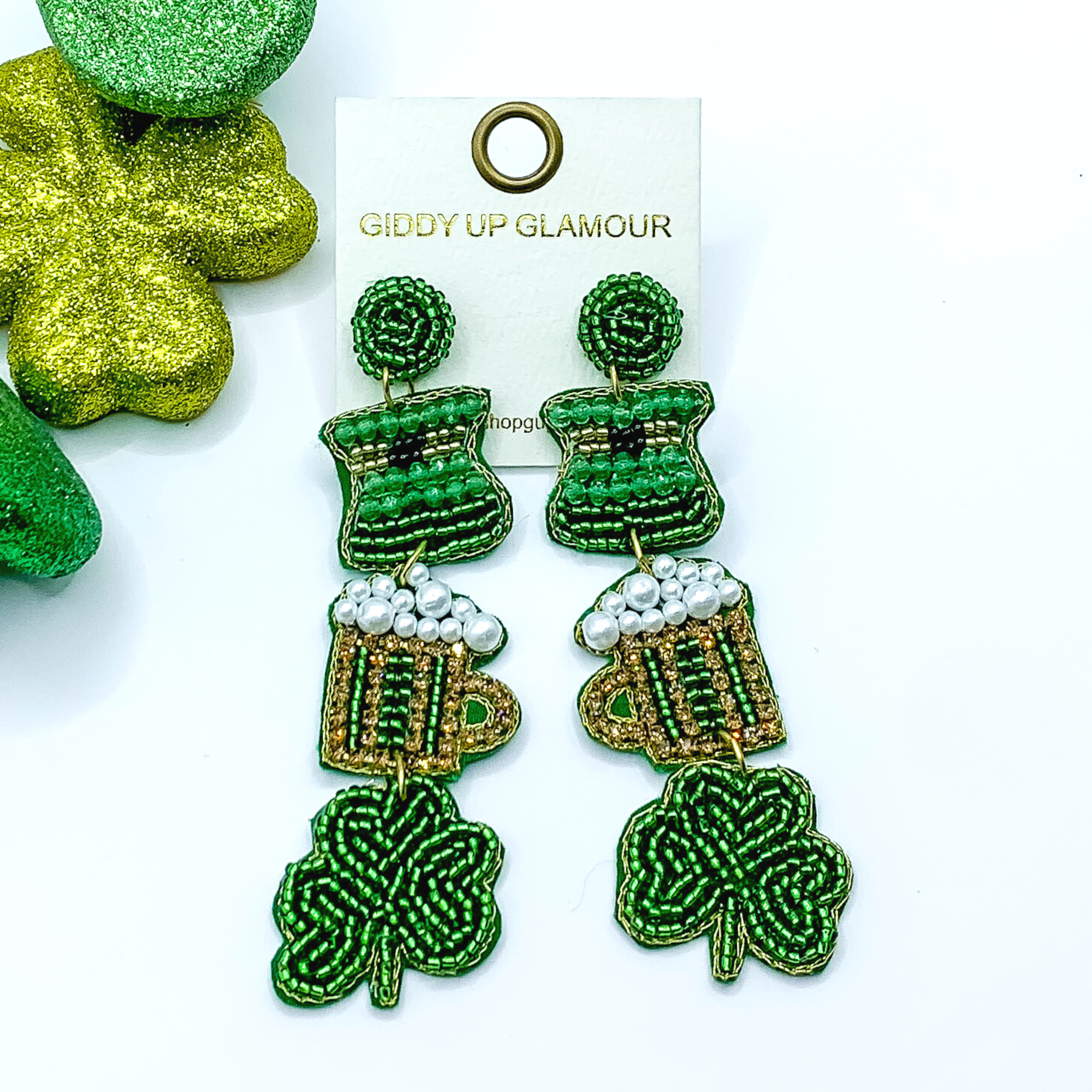 Green beaded drop earrings. These earrings include a green pot, a crystal beaded mug, and beaded clover. These earrings are pictured on a white background with green decor in the top left corner. 