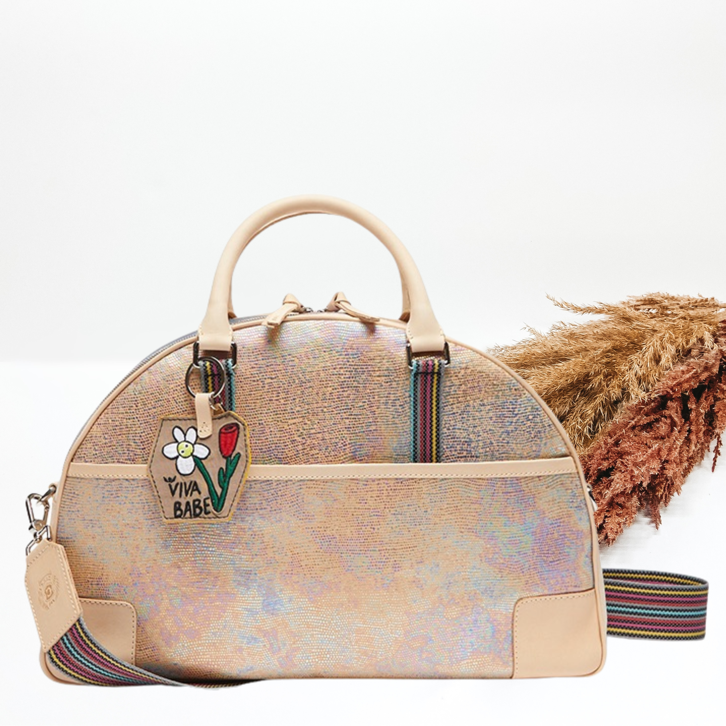 Pictured is a dome shaped bag with light tan short handles and a striped strap. This bag is a multicolor iridescent print with light tan accents and a charm on one of the handles. This bag is pictured on a white background with pompous grass on the right side of the bag. 
