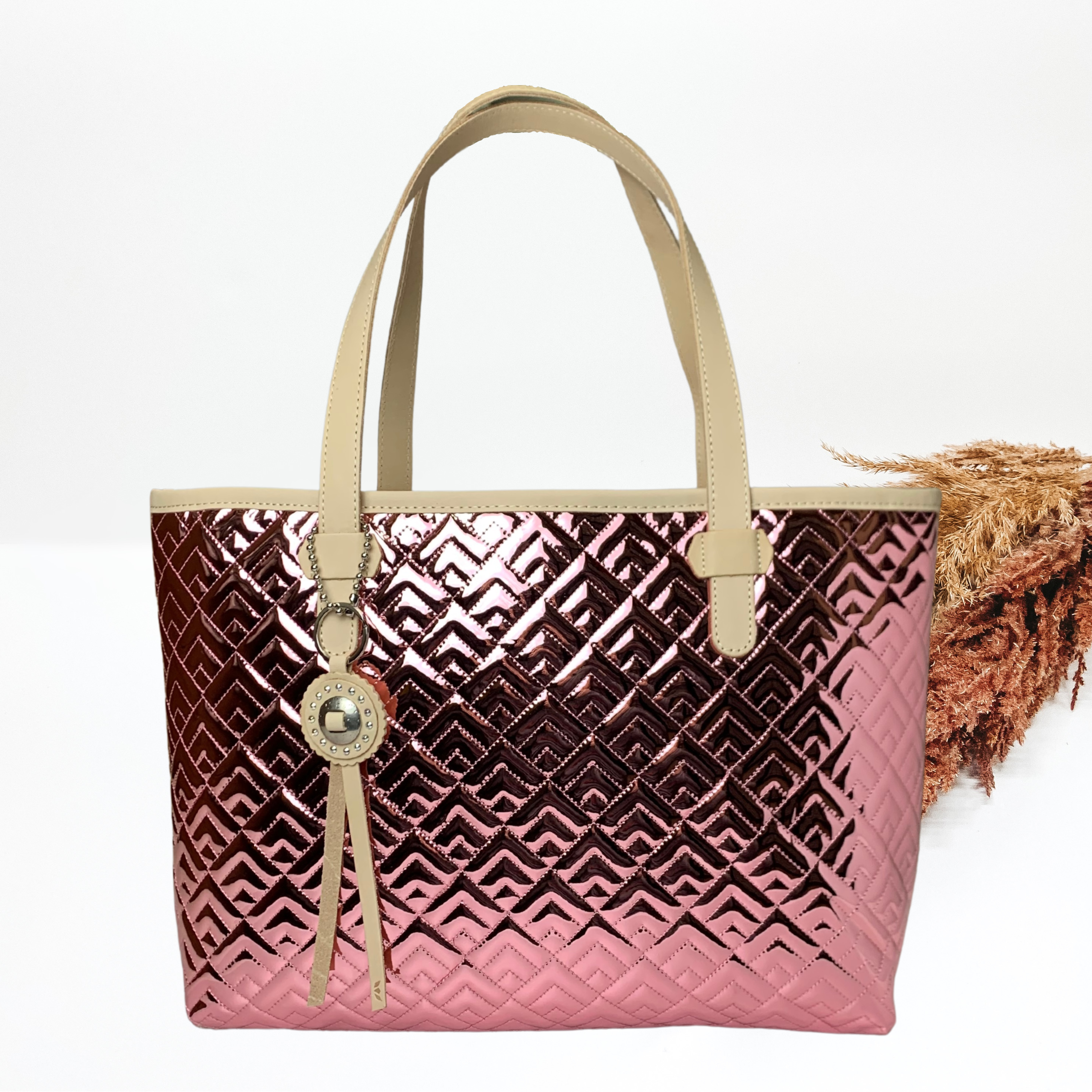 Pink metalli tote bag with a stitched pattern. This bag has light tan handles, a light tan tassel, and is a rectangle shape. This tote is pictured in front of pompous grass on a white background.