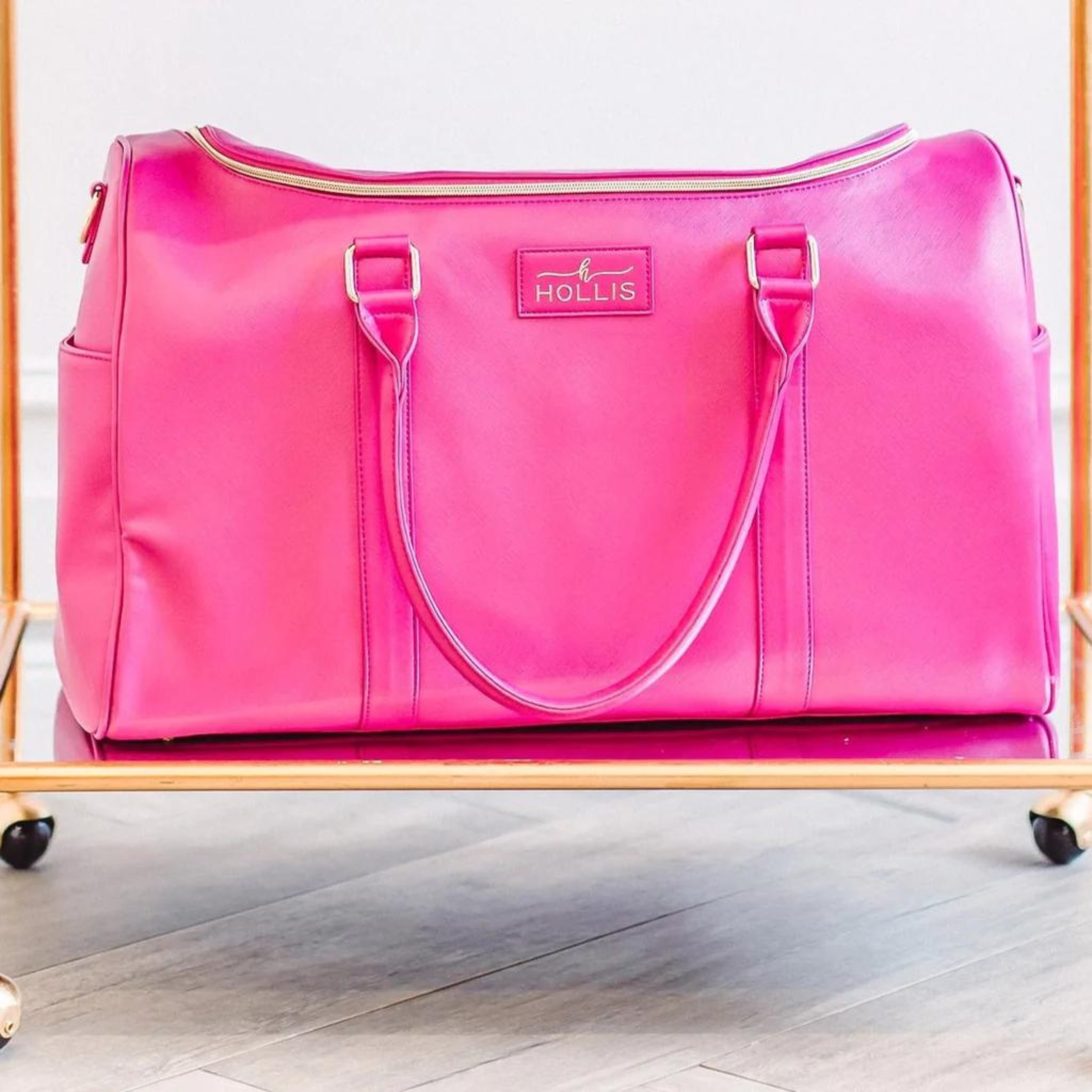 Hot pink duffel bag with hot pink handles. This bag has a gold top zipper and pocket on each side. This bag is pictured on a gold cart in front of a light grey wall. 