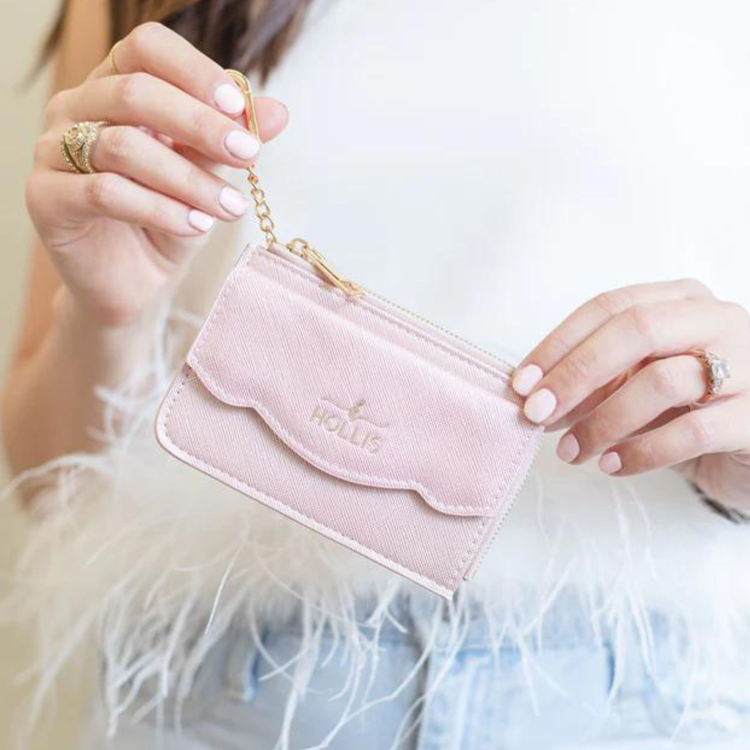 Pictured is light pink card holder with a gold key ring. This card holder also has the brand "HOLLIS" in the center. This card holder is pictured by someone holding it in front of a white feather top. 