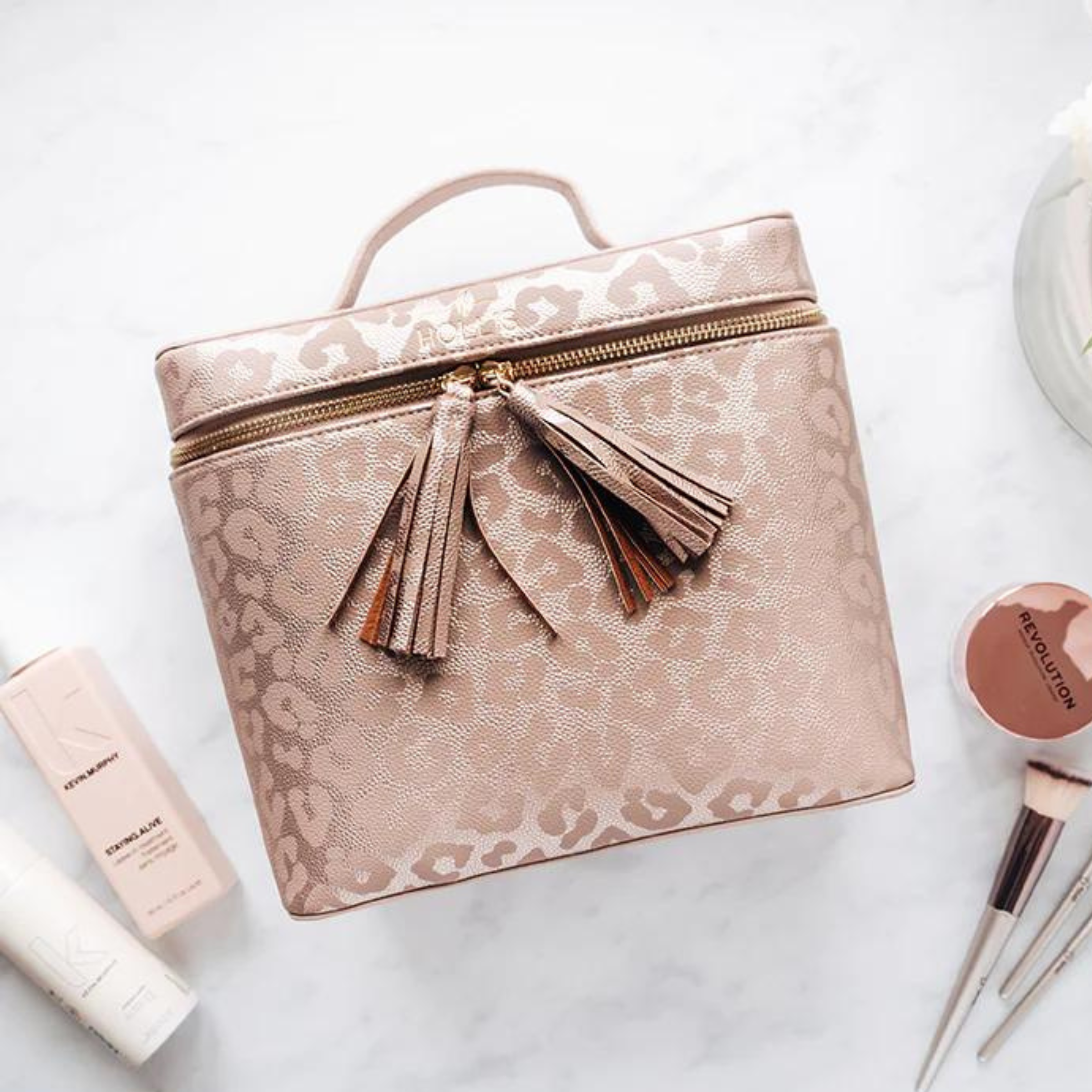 Pictured is a rectangle pink and bronze bag with a leopard print design. This bag includes an all around zipper with tassel pulls and a top handle. This bag is pictured on a white background with beauty products at the bottom of the picture. 