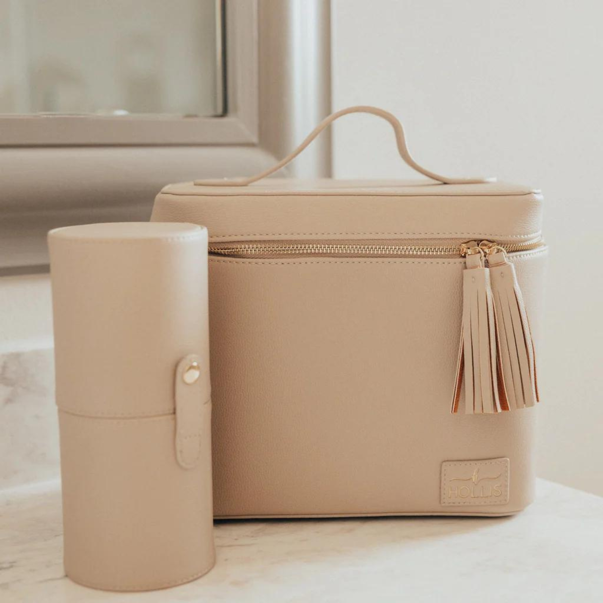 Pictured is a rectangle nude bag that includes an all around zipper with tassel pulls and a top handle. This picture includes a nude cylinder brush holder and is all pictured on a off white background.  