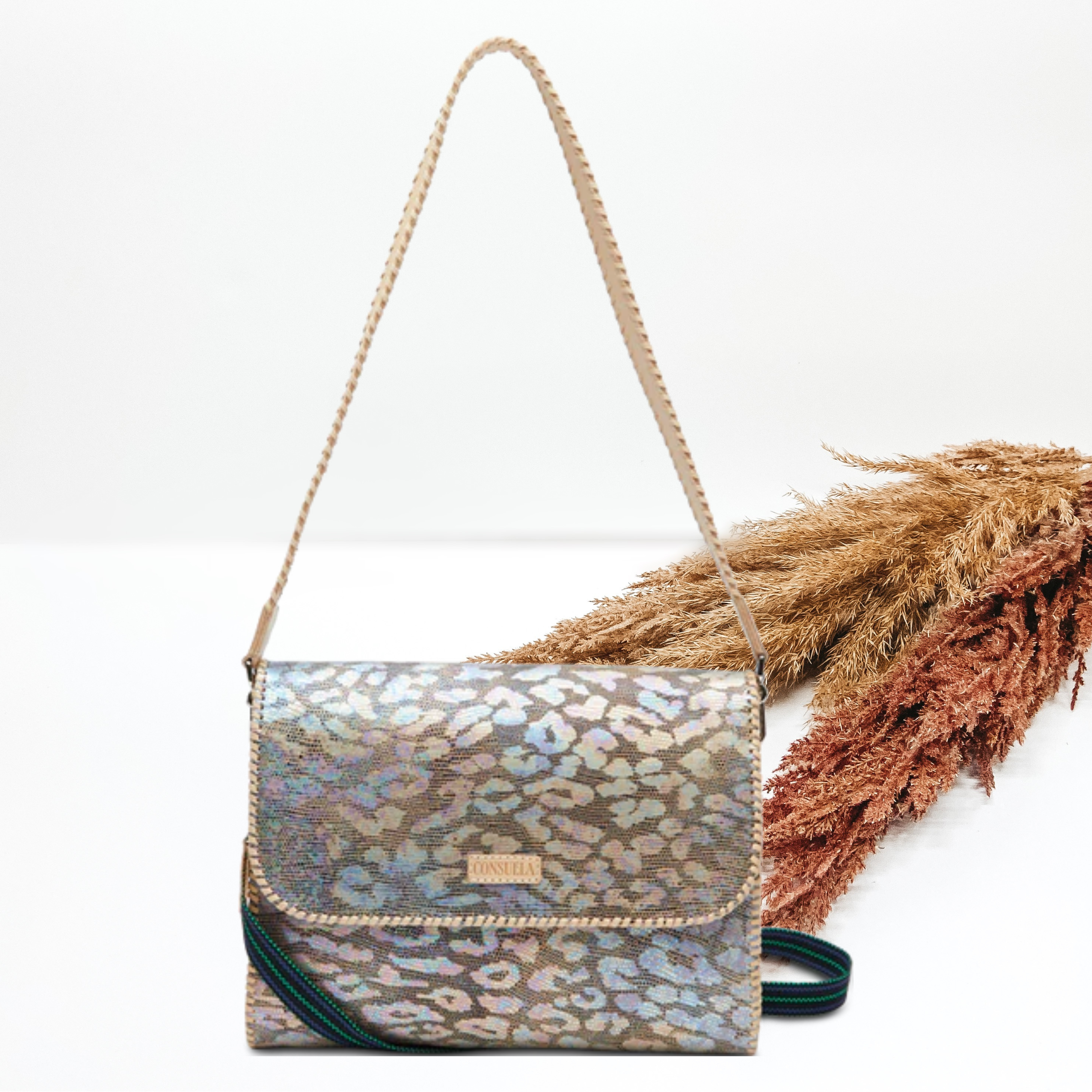 Pictured is a rectangle purse with a light tan strap and a stitched, striped strap. This bag has an iridescent, multicolor leopard print design with a light tan outline stitching. This bag is pictured on a white background with pompous grass behind it. 