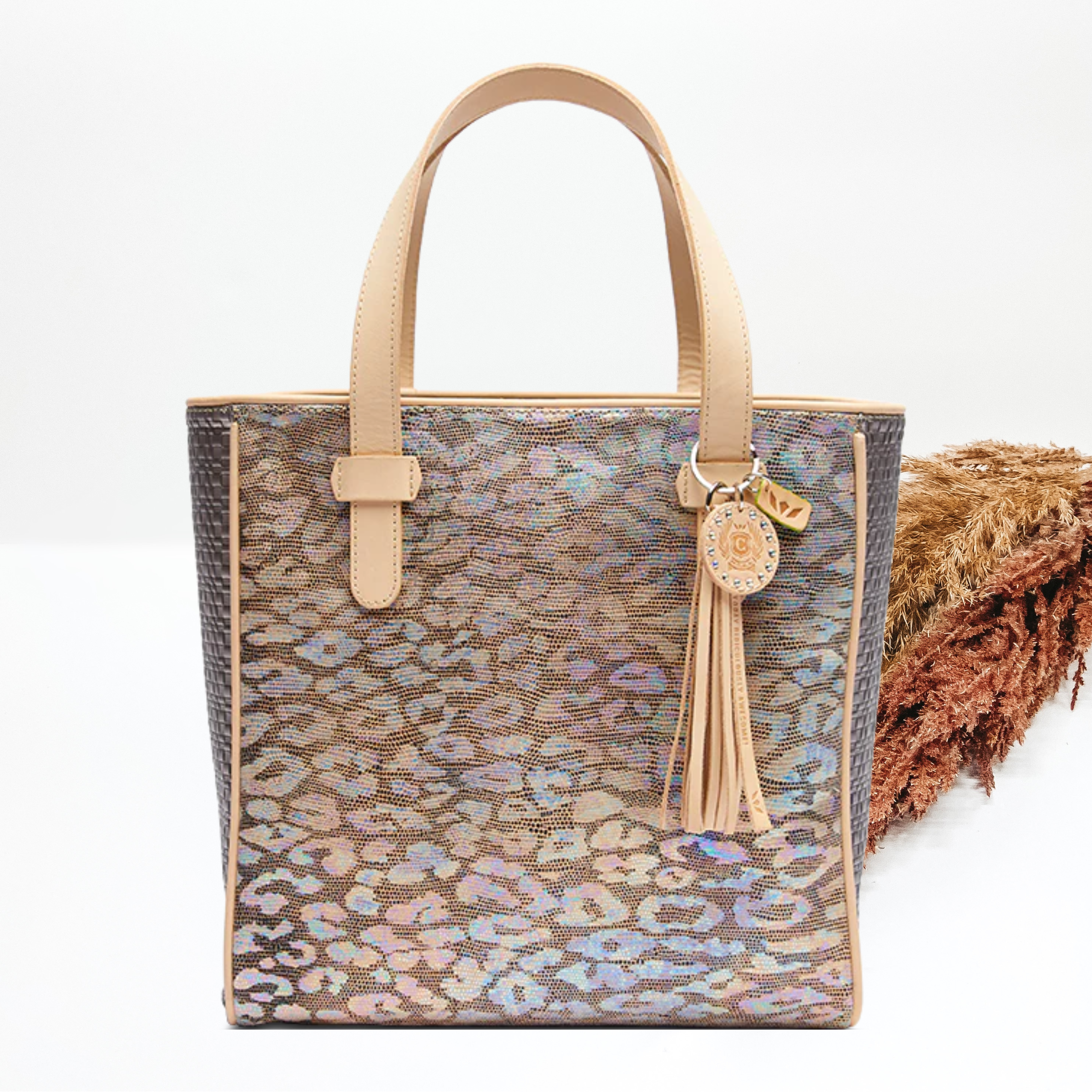 Pictured is a rectangle tote with light tan shoulder straps and a iridescent, multicolor leopard print design. This tote also includes a light tan tassel and key chain. This tote is pictured on a white background with pompous grass behind it. 