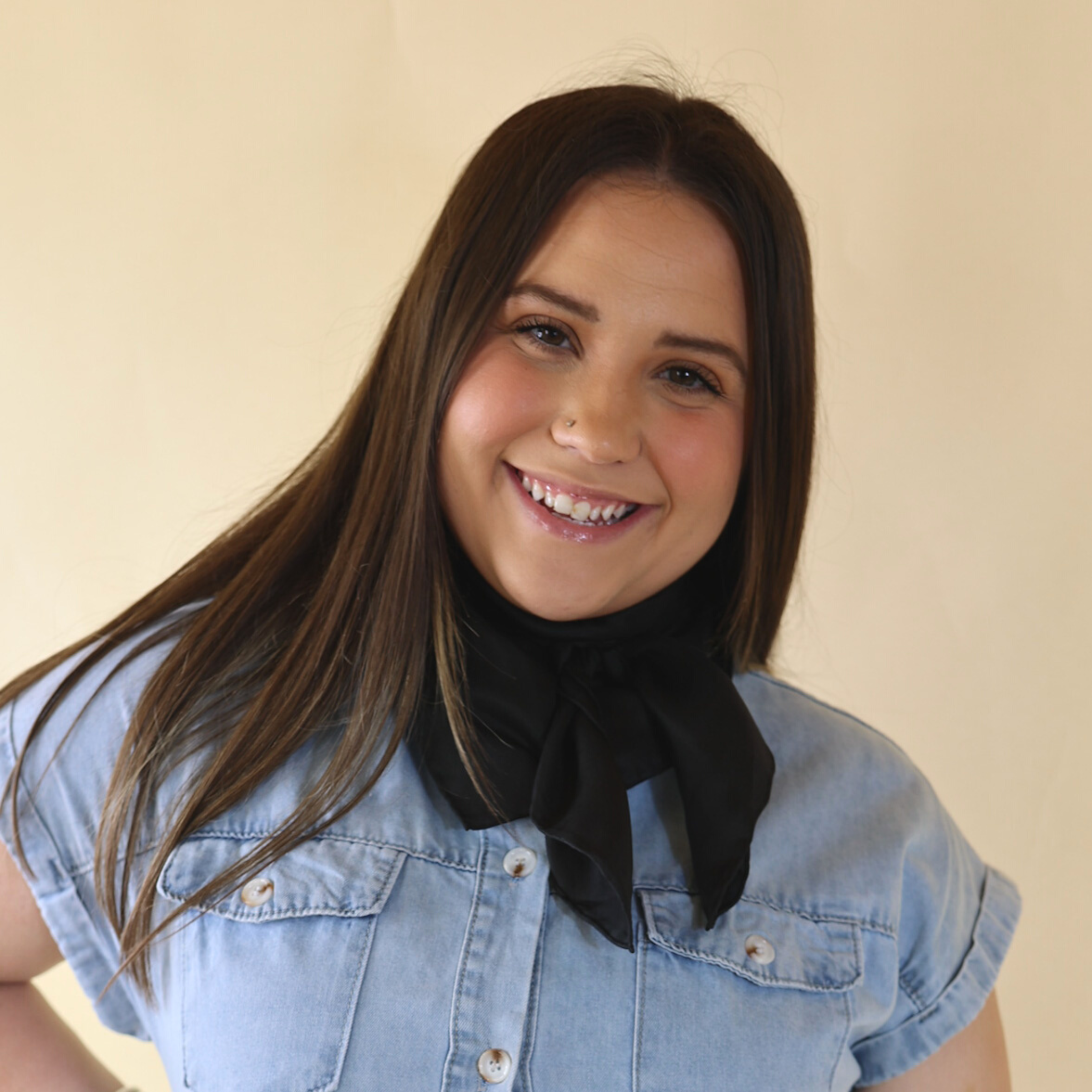 Brunette model wearing a short sleeve, denim button up with a solid black scarf tied around her neck. This model is pictured in front of a beige background. 