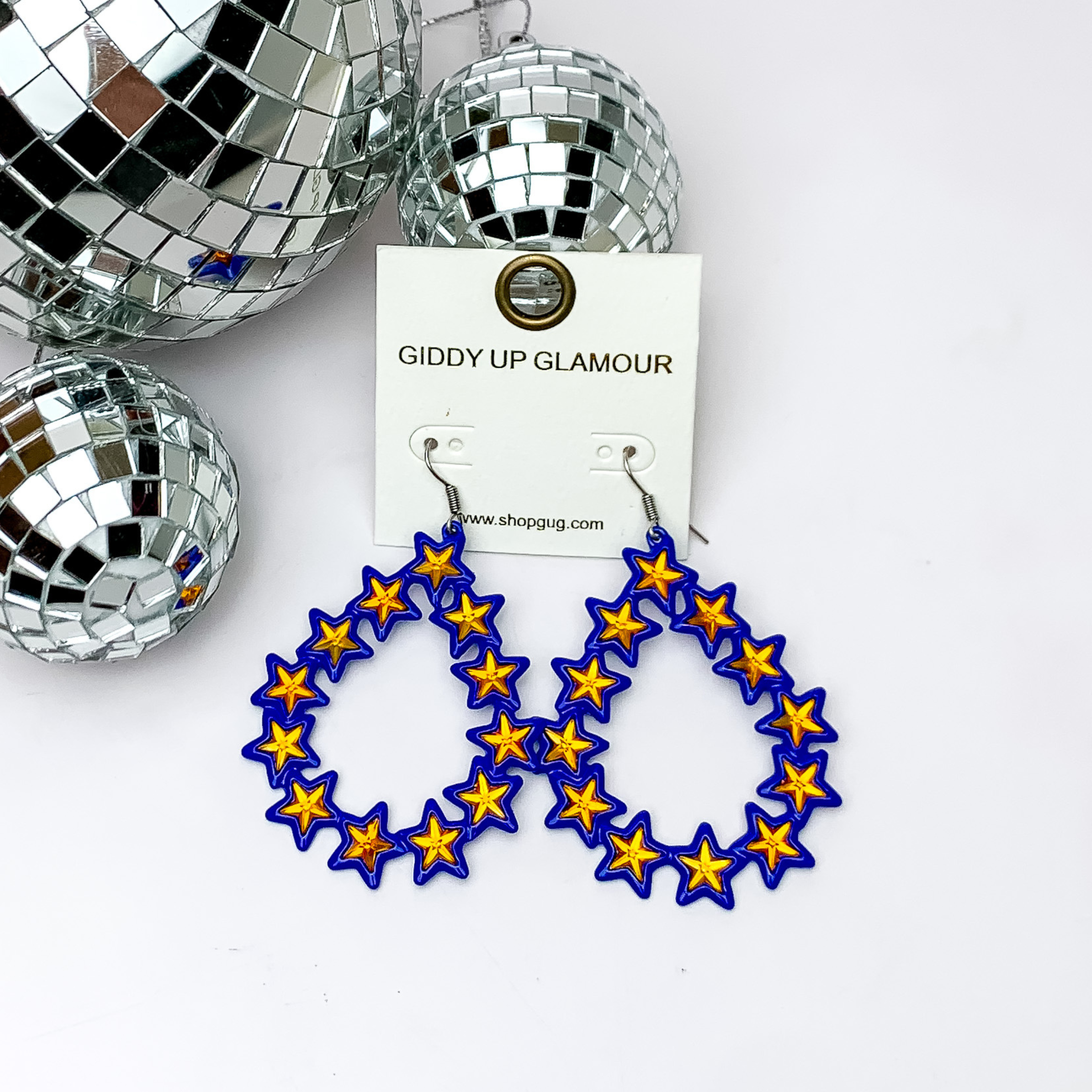 Blue, star linked teardrop earrings with orange centers on a white earring holder. These earrings are pictured on a white background with disco balls in the top left corner. 