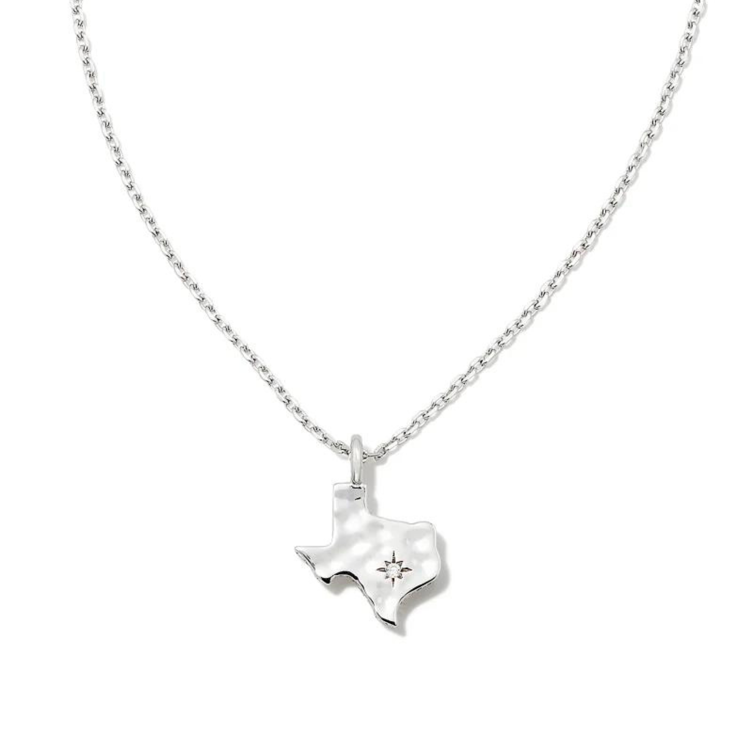 Pictured on a white background is a silver chain necklace with a silver texas shaped charm that has a hammered texture and a tiny crystal on it. 
