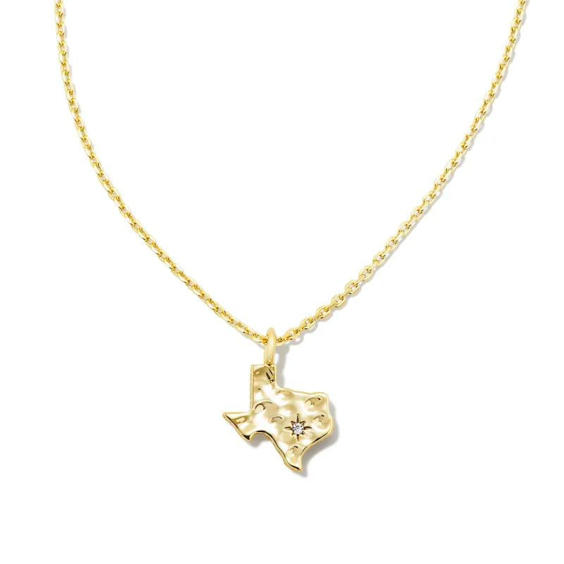 Pictured on a white background is a gold chain necklace with a gold texas shaped charm that has a hammered texture and a tiny crystal on it. 