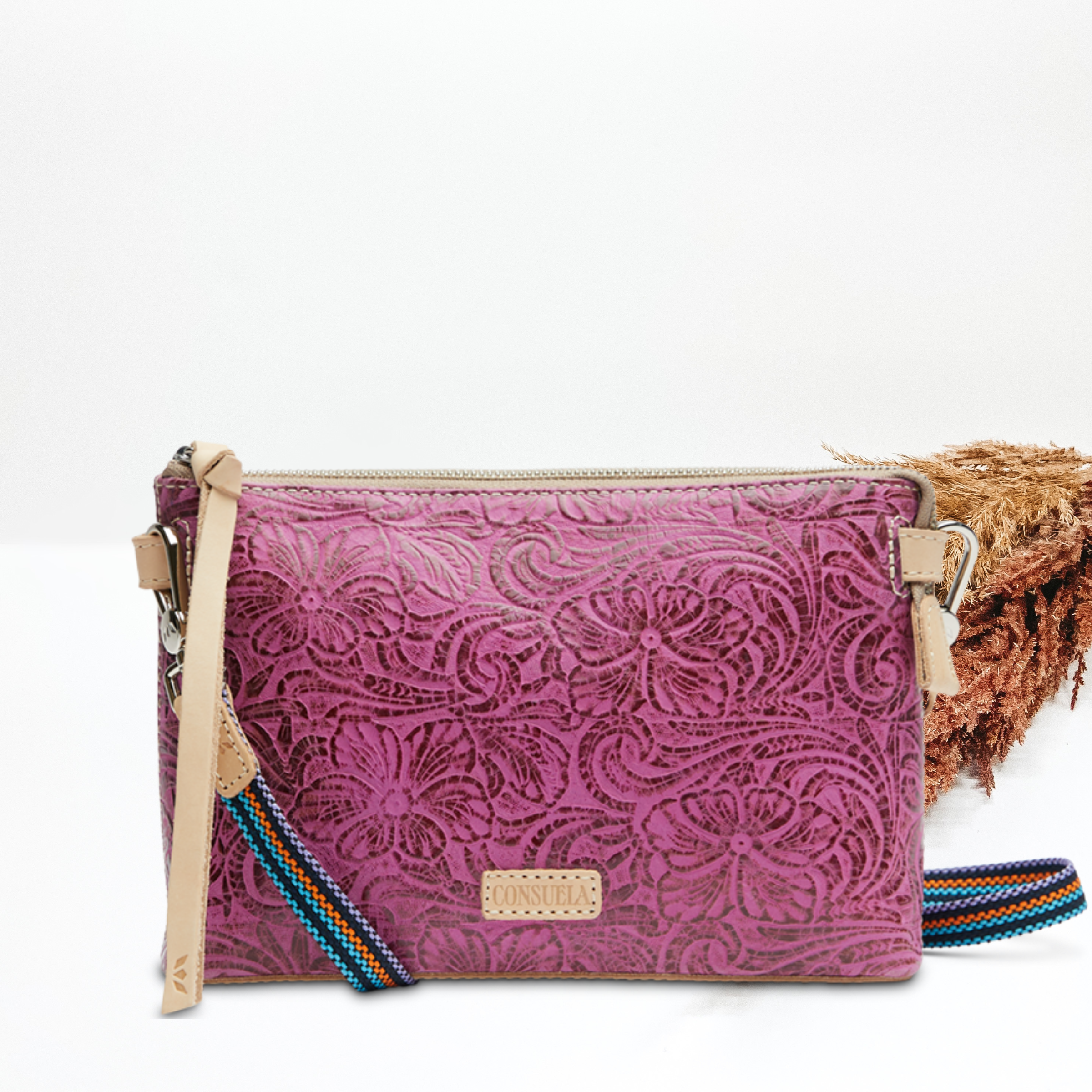 Pictured on a white background is a fuchsia, tooled leather print midtown crossbody. This purse includes a top zipper with a light tan leather tassel and a striped purse strap.   