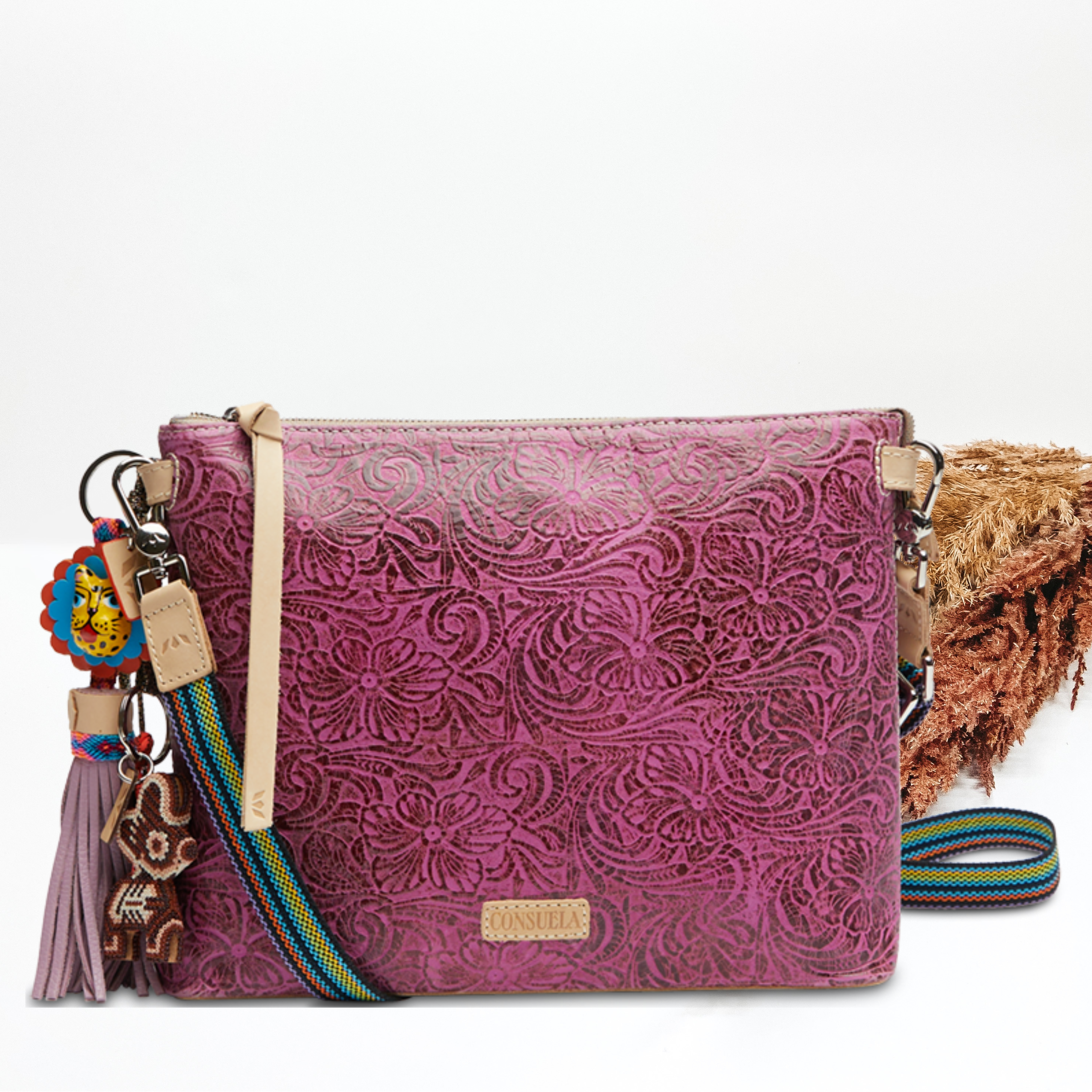 Pictured on a white background is a fuchsia, tooled leather print midtown crossbody. This purse includes a top zipper with a light tan leather tassel, a striped purse strap, and two multicolor purse charms.   